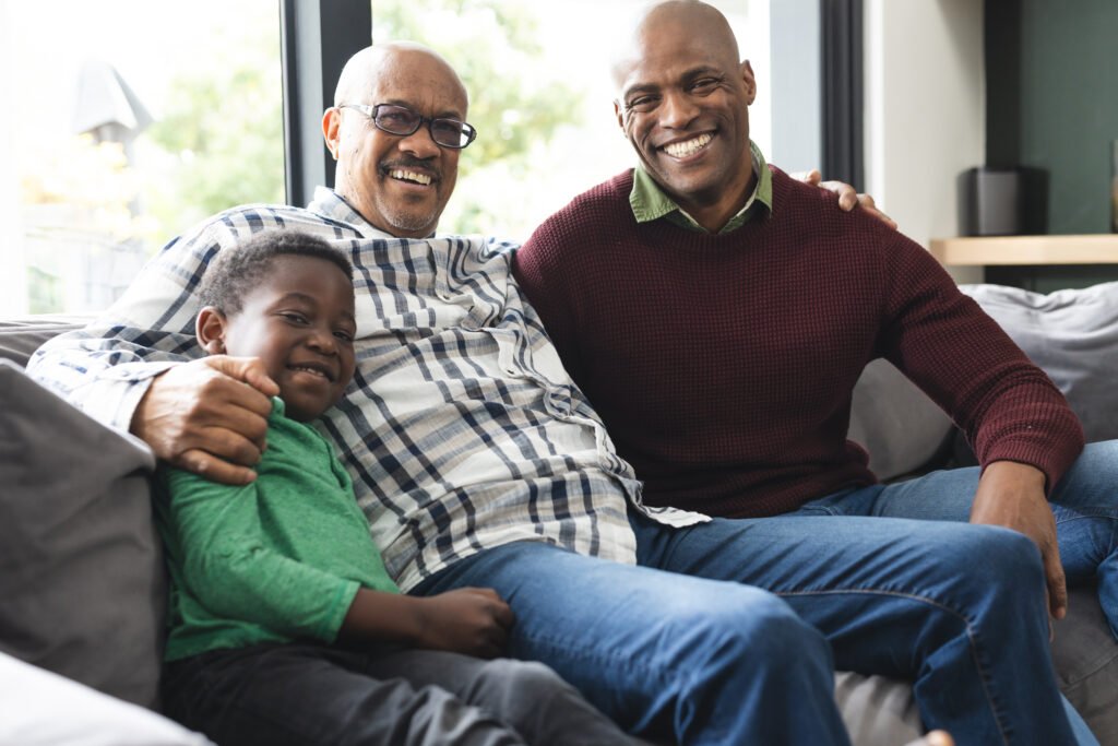 Happy african american father, son and grandfather sitting on couch,laughing and embracing. Childhood, home, family, togetherness, domestic life and lifestyle, unaltered.